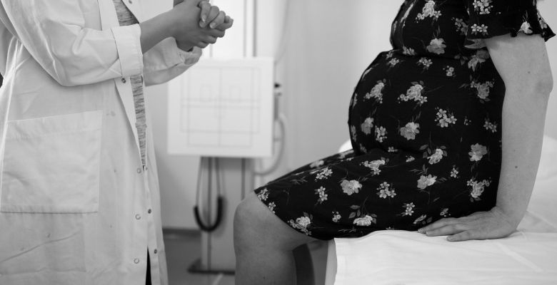 a pregnant woman in a patterned dress sits on the edge of a hospital bed. a female doctor is speaking to her, with hands clasped.