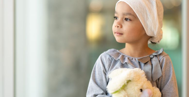 A beautiful little girl with cancer takes a break from treatment. She is standing near a large bay of windows in the hospital's corridor. The girl is wearing a headscarf and is holding a stuffed rabbit toy. She is looking out the window with a peaceful expression.