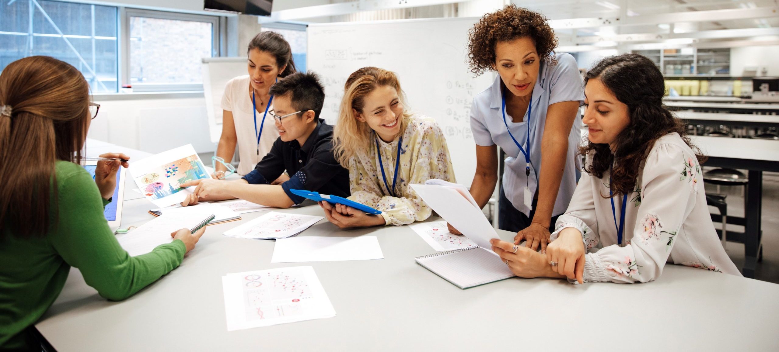 Teacher with a group of university students, in a laboratory classroom. The instructor is considering one of the students work, the mood is light hearted and positive. Other classmates are discussing things with each other. This is a realistic teaching scenario, with candid expressions. This is a multi-ethnic group of women. In the background there is a white board with mathematical formula written on it. All ladies are wearing id tags.