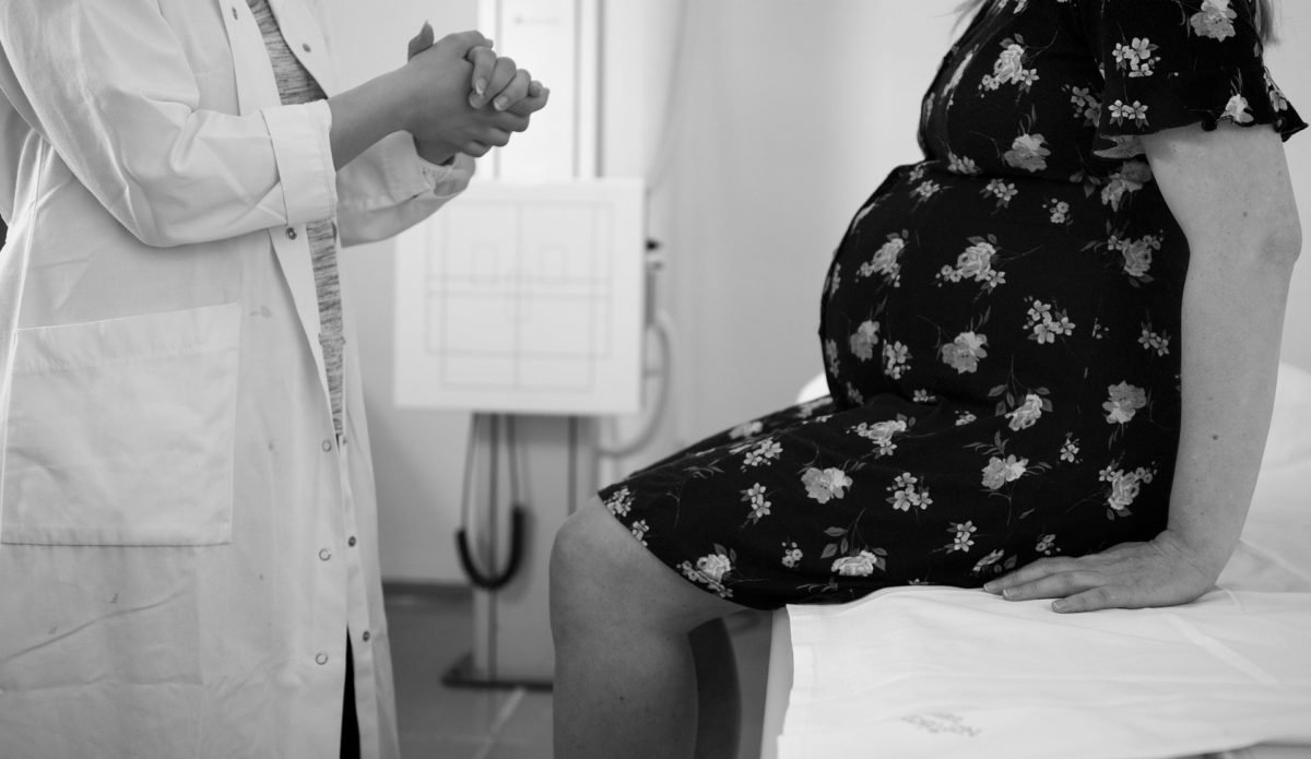 a pregnant woman in a patterned dress sits on the edge of a hospital bed. a female doctor is speaking to her, with hands clasped.