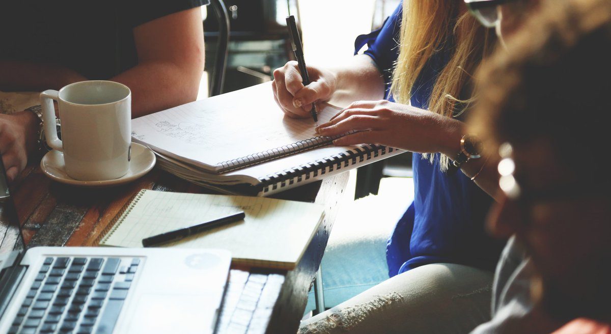 a group of adults in a meeting. a woman is writing in a spiral bound note book.