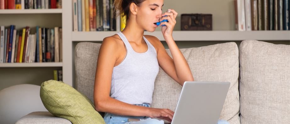 young girl having an online asthma appointment and using an inhaler sitting on a sofa at home with laptop